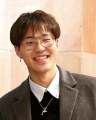 Shuzhe (Jerry) Guan with short brown hair, wearing a shirt, sweater, and grey blazer, in front of peach-colored stone building 