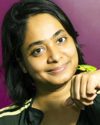 Sonali Garg with long black hair, a bright green frog sitting on the back of her hand, in front of a fuschia background