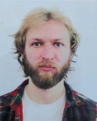 Taylor Brock-Fisher with light hair and dark beard, wearing white shirt and plaid flannel, in front of white background