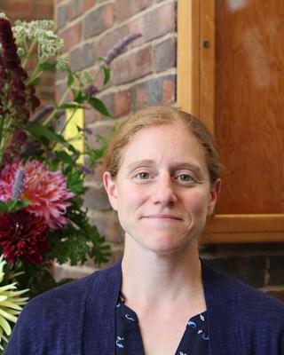 Tracy Barbaro - light red hair pulled back, blue shirt, in front of brick wall and flowers indoors