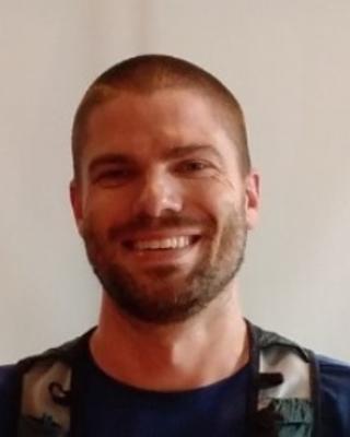 Tyler Coverdale with short brown hair and beard, wearing a dark shirt and backpack, in front of a light grey background
