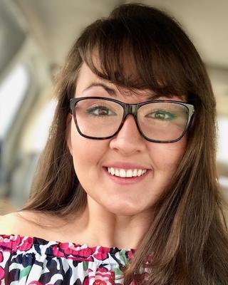 Valeria Schmidt with long brown hair and bangs, wearing glasses and colorful patterned shirt, windows in background