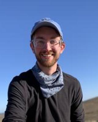 Walker Weyland with a short brown beard, wearing glasses, a blue hat and scarf, and black top, with blue sky and brown hill in background