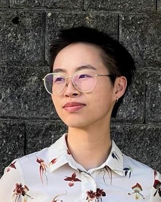 Yi Lin Zhou with short dark hair, wearing glasses and a light top with dark orchid pattern, in front of grey brick wall