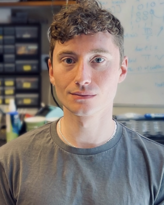 Tom Alisch with curly dark blonde hair, wearing a grey top, in front of blurred lab background