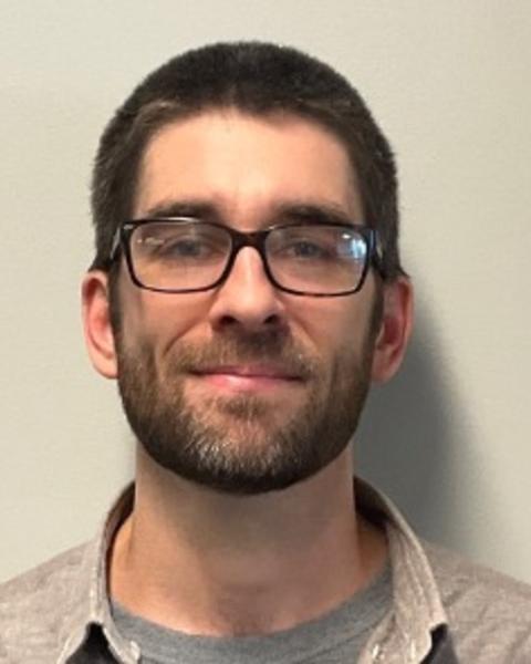 Peter D Wiggan with short brown hair and beard, grey t-shirt, grey button down, light grey background