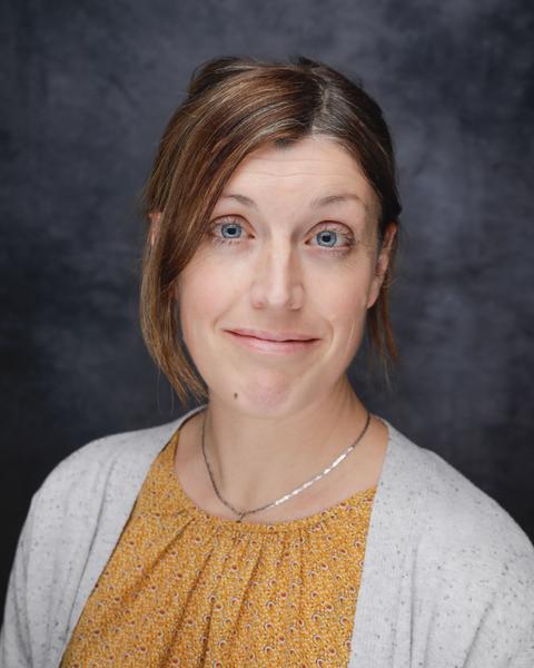 Elsa Goerig with brown hair pulled back, wearing a patterend yellow shirt and white cardigan, in front of a dark grey background