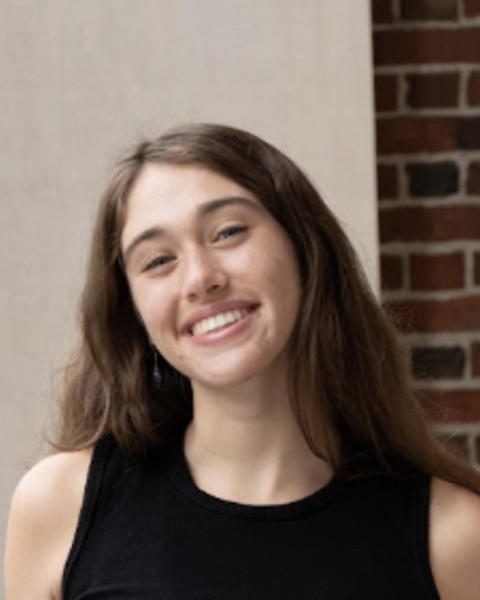 Beatrice Youd with long brown hair and black sleeveless top, in front of cream wall and brick column
