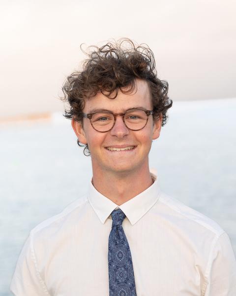 Max Laubstein with short curly brown hair, wearing glasses and a white shirt and blue tie, with the ocean in the background