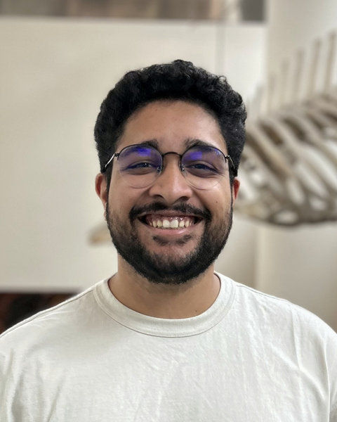 Akshay Kan with short black hair and beard, wearing glasses and a white shirt, with fossil in background