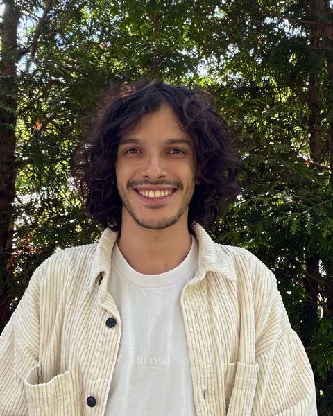 Lucas Funaro with long dark wavy hair, a white shirt and cream corduroy shirt, in front of green trees