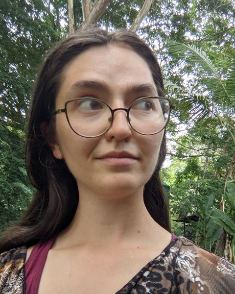 Portrait of Sarah Dendy. Woman with long dark hair wearing glasses and standing outside