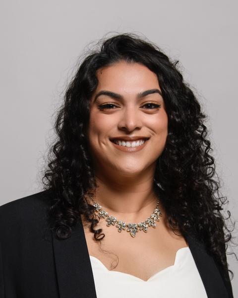 Amarilis Castro smiling, with long curly black hair, black blazer, white scalloped shirt, grey background