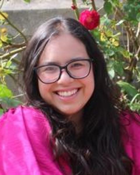 Andrea Loya Castillo with long dark hair, glasses, and a bright pink top, in front of dark pink rose bushes