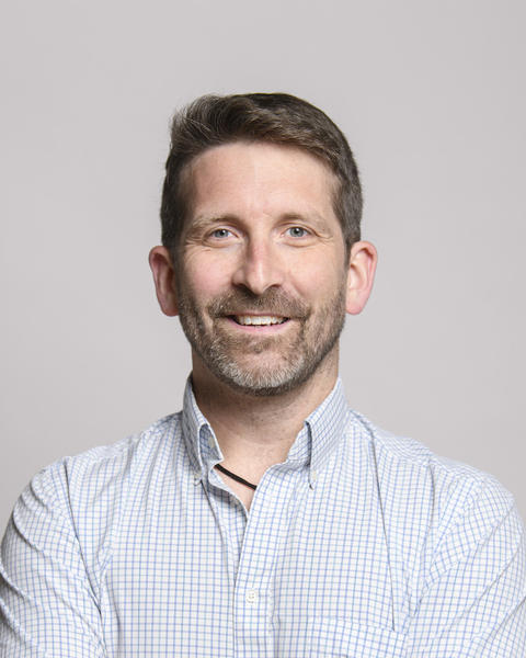 Benton Taylor with dark hair and a beard wearing a white shirt, in front of grey background