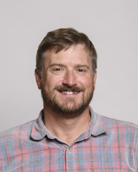 Christian Flynn smiling, short brown hair and beard, red and blue plaid shirt, grey background