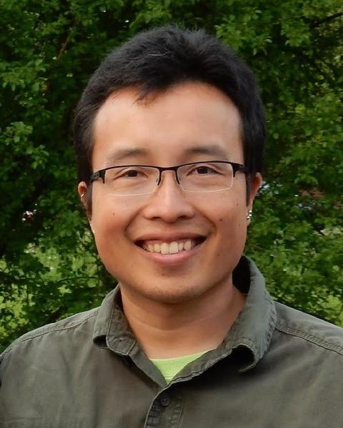 Evan Hoki with short dark hair, glasses, and a muted green shirt, in front of green leafy trees