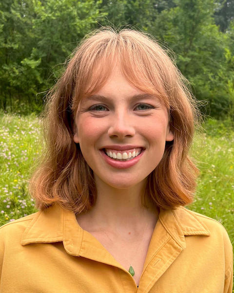 Eve Farrell with medium length red hair and bangs, wearing a yellow shirt, in front of trees and a flowering meadow