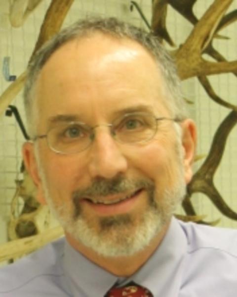 James Hanken, with short grey hair and beard, wearing glasses and smiling in front of a wall of antlers
