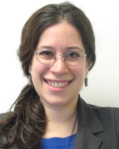 Joanna Wolfe with long dark hair pulled back, wearing glasses, a blue top, and a dark blazer, in front of a white background
