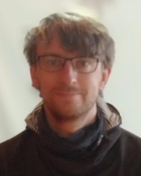 Jonas Geburzi with short brown hair, wearing glasses and a dark coat, in front of a beige background
