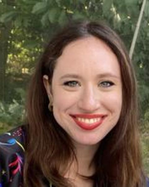 Lily Shapiro with long brown hair, black shirt with bright pattern, in front of leafy trees