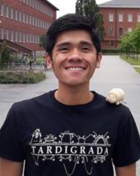 Marc Mapalo with short dark hair, black tshirt with "tardigrade" on it, campus behind him