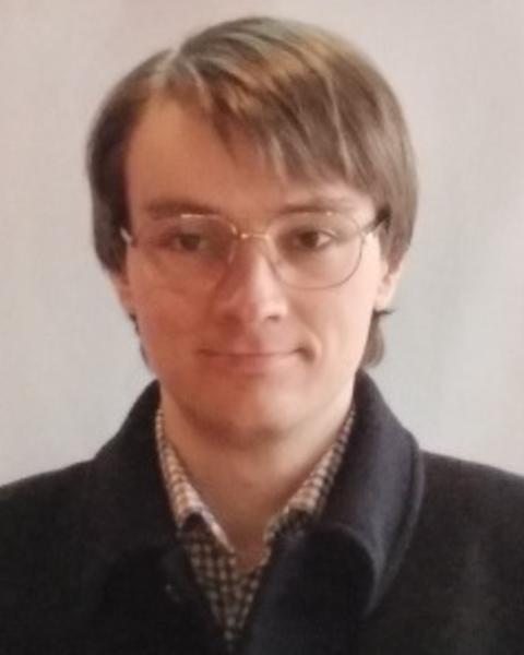 Peter Bishop with light brown hair, wire glasses, dark coat, in front of grey background