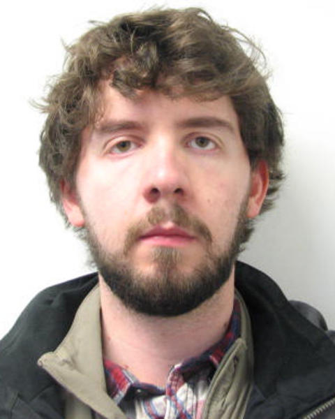 Robert Brocklehurst with short, curly brown hair and beard, wearing layered shirts and dark coat, in front of white background