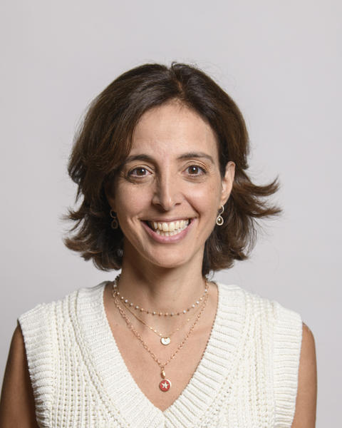 Sarine Der Kaloustian, smiling, with short brown hair, cream sleeveless sweater, grey background