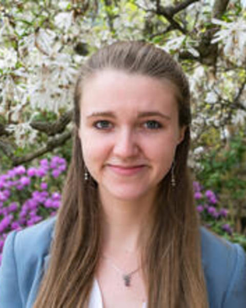 Sophie Everbach with long brown hair and a blue blazer, in front of white and purple flowering tree and shrubs