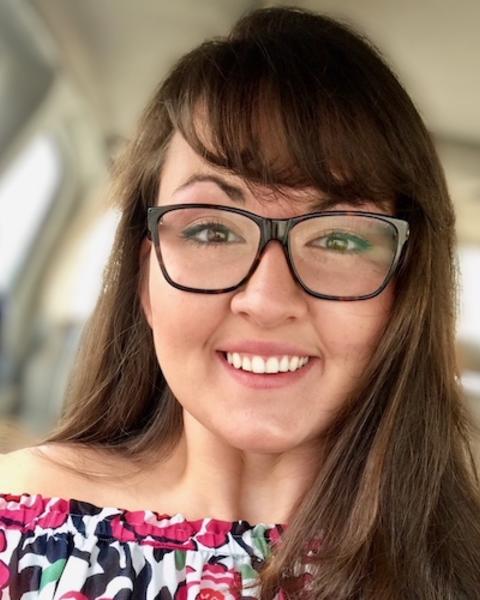 Valeria Schmidt with long brown hair and bangs, wearing glasses and colorful patterned shirt, windows in background
