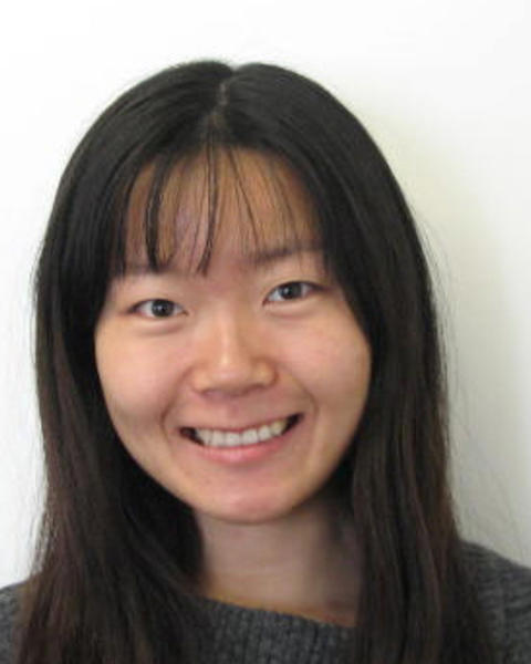 Yujing Yan with long dark hair and bangs, wearing a grey sweater, in front of a white background