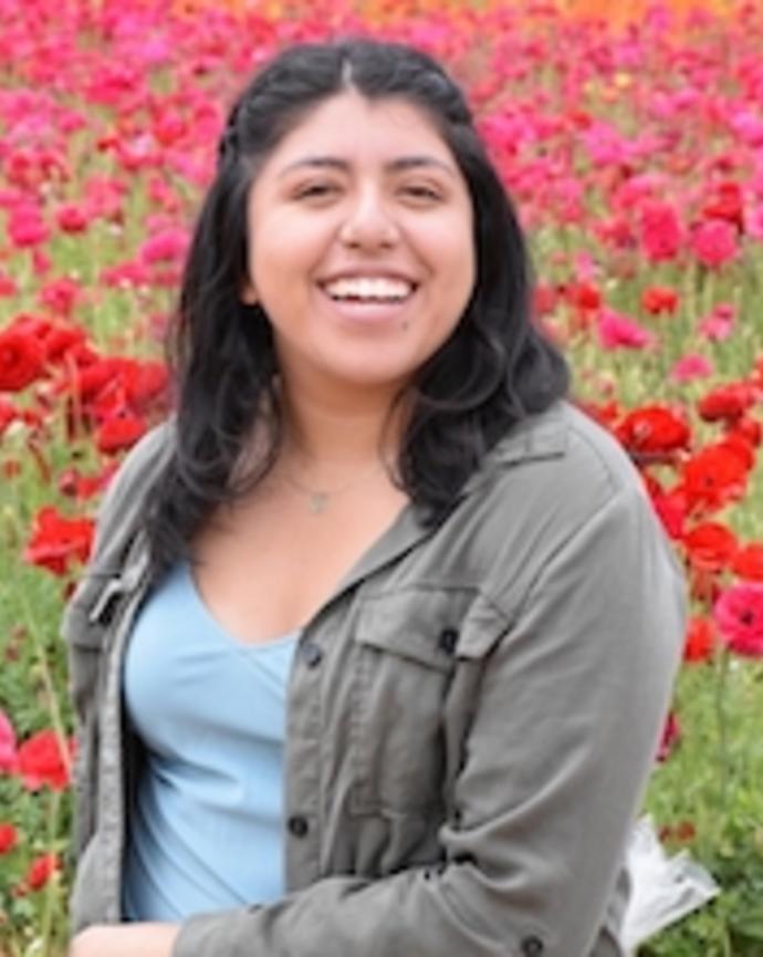 Adriana Aguilar-Maldonado with long dark hair, blue top and grey jacket, in a field of red flowers