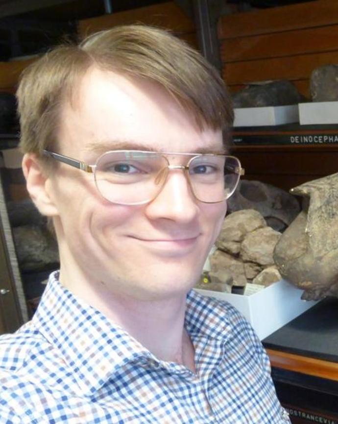 Portrait of postdoc Peter Bishop standing in front of a fossil wearing a plaid shirt and glasses