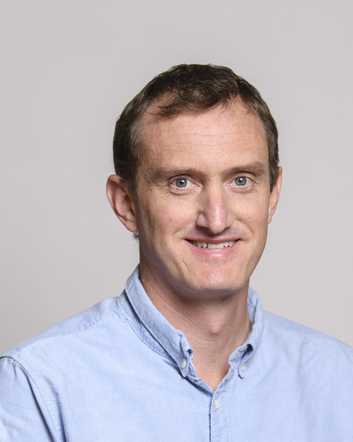 Andrew Davies with short dark hair and blue button up shirt, in front of light grey background