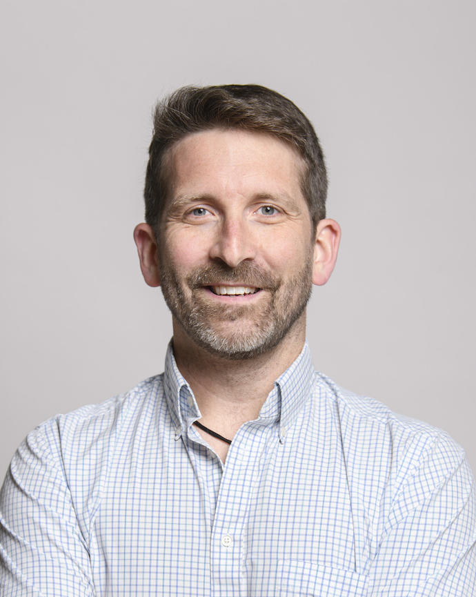 Benton Taylor with dark hair and a beard wearing a white shirt, in front of grey background