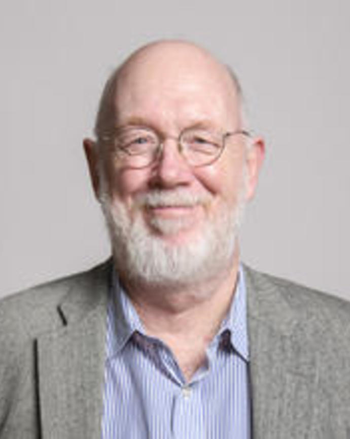 David Haig with gray hair, a beard, wearing glasses and a suit, in front of grey background