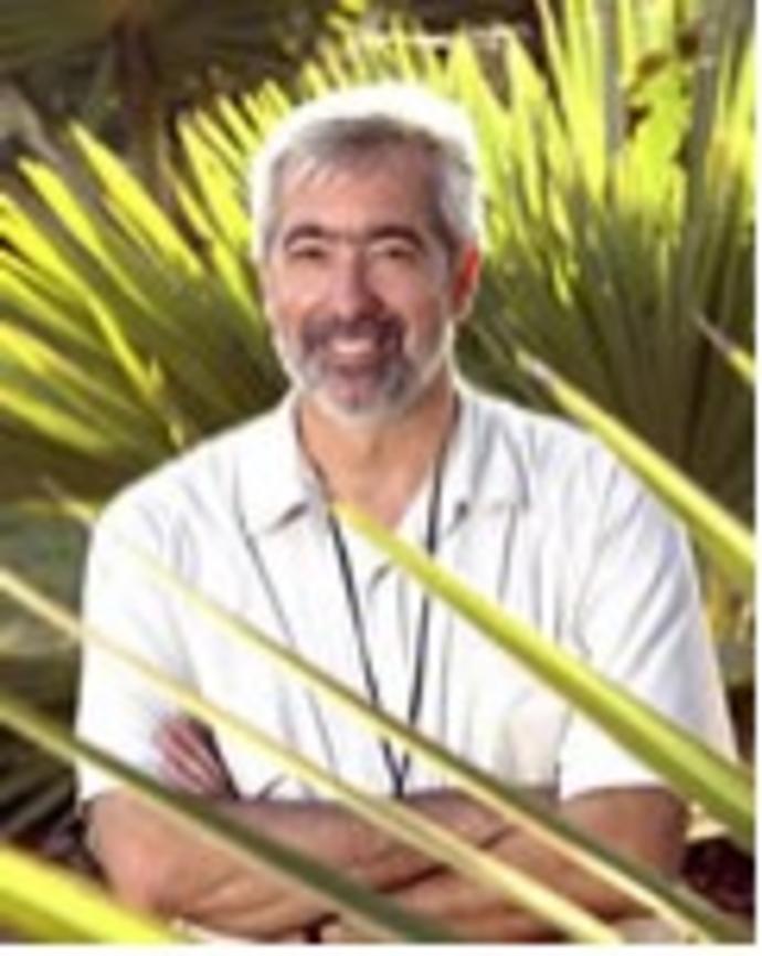 Brian D. Farrell with grey beard and hair, wearing white short sleeve button down, with spike green leaves behind him