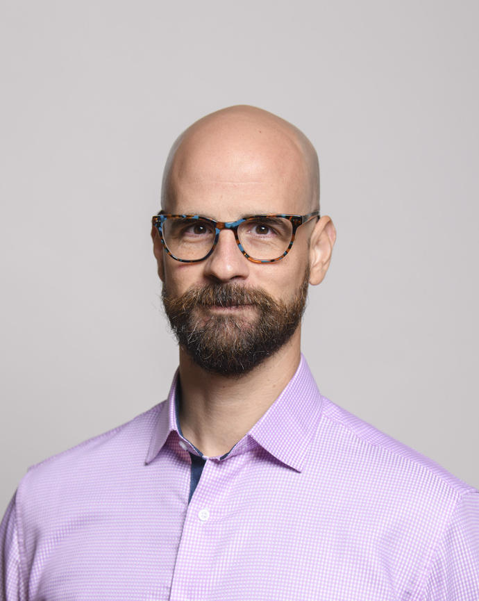 Javier Ortega-Hernandez wearing colorful glasses, a pink check shirt, and with a short dark beard, in front of grey background