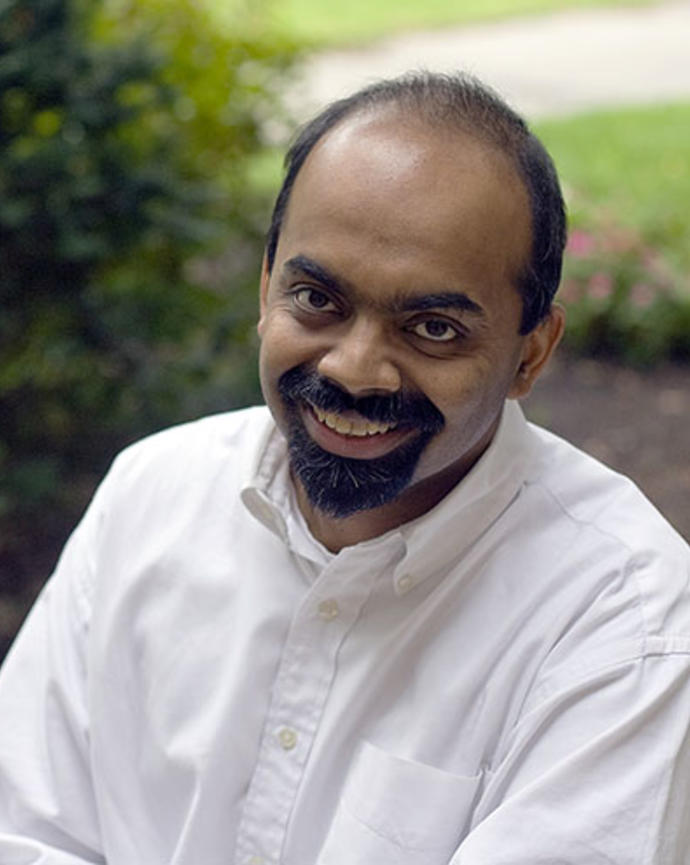 L. Mahadevan with short dark hair and beard, wearing white button down shirt, with grass and shrubs behind him