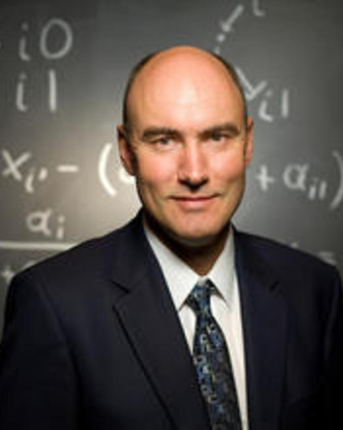 Martin A. Nowak wearing a suit and tie, in front of a chalkboard with equations on it