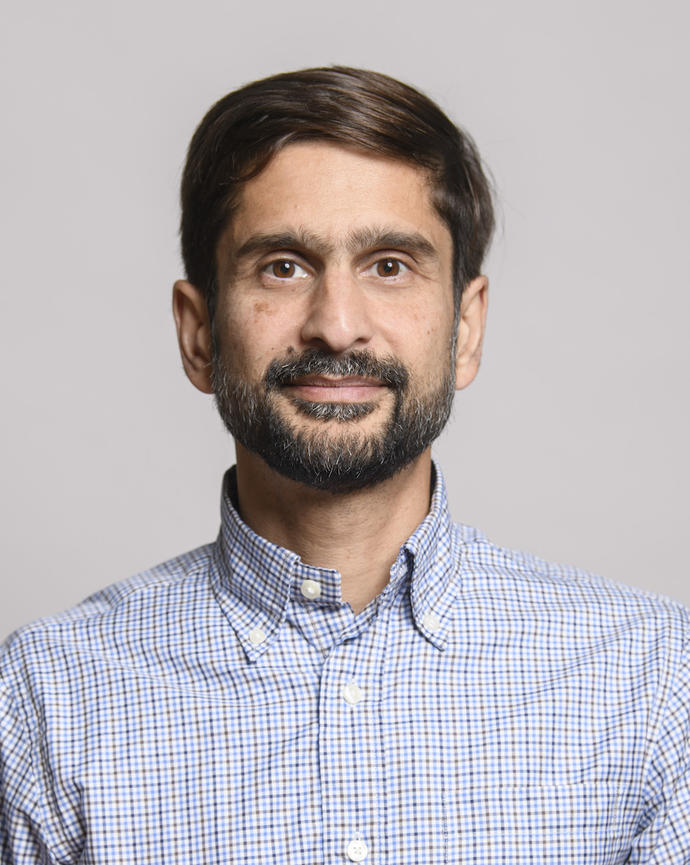 Michael Desai with short dark hair and beard, wearing a blue check button down, in front of grey background