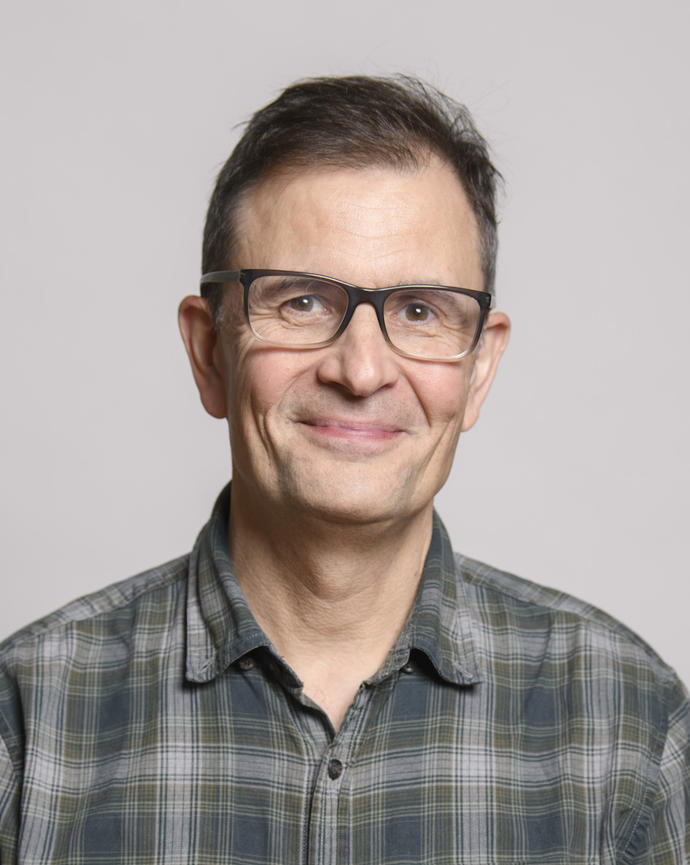 Paul Moorcroft smiling, with short hair, plaid button down, and glasses, in front of grey background