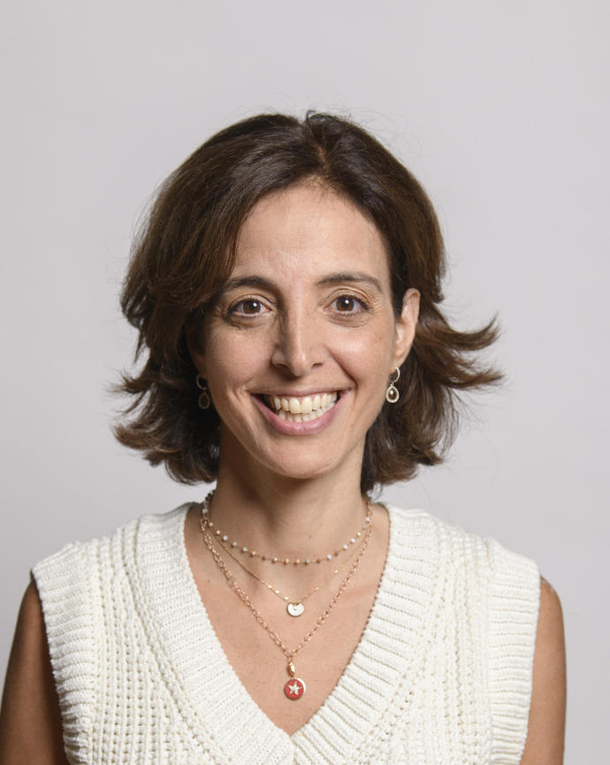 Sarine Der Kaloustian, smiling, with short brown hair, cream sleeveless sweater, grey background