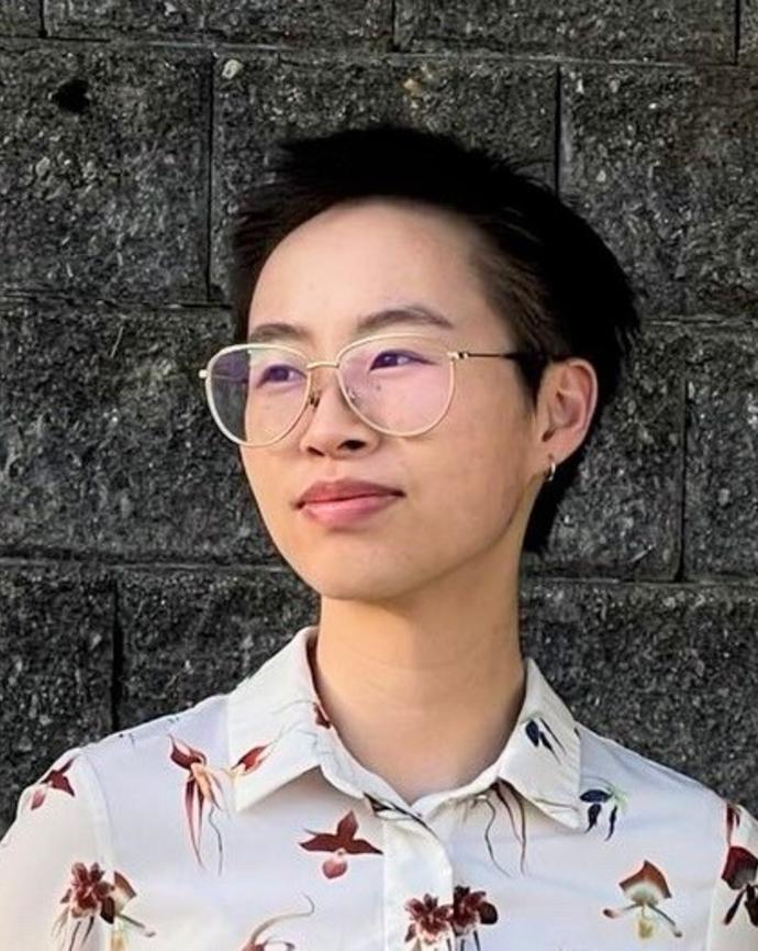 Yi Lin Zhou with short dark hair, wearing glasses and a light top with dark orchid pattern, in front of grey brick wall
