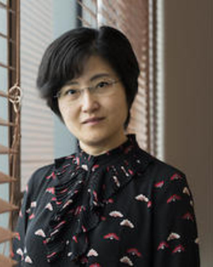 Yun Zhang with short black hair, glasses, and black patterned shirt, standing near window