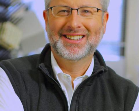 Man with beard and glasses wearing black vest and white shirt smiling