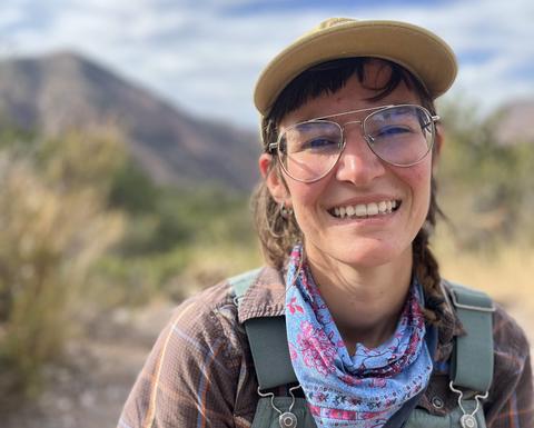 Graduate student Juli Mayorga standing in front of a mountain wearing overalls, hat and glasses and smiling