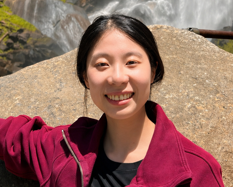Portrait of PhD candidate Jingting Liang standing outside by a waterfall wearing a red sweater